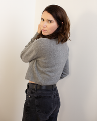 A woman stands in front of a wall, elegantly dressed in the Autumn Cashmere Cropped Cardigan in Cement.