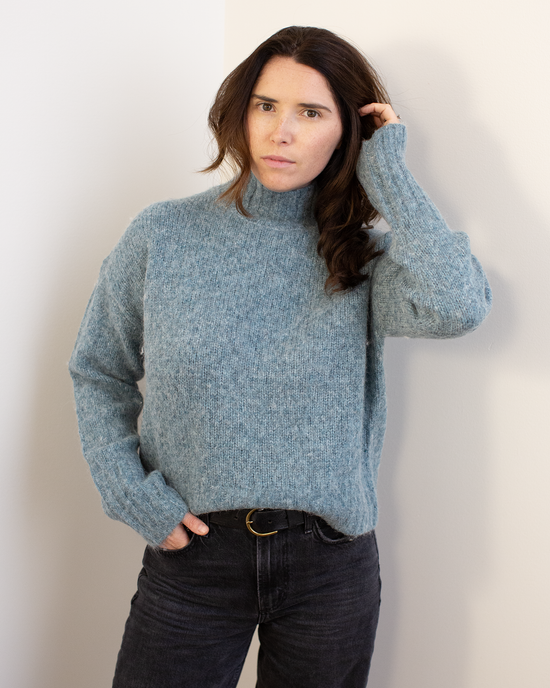 A person with long brown hair wears the Hartford Myac Turtleneck Sweater in Jade and black jeans, standing against a white wall with one hand in their pocket and the other touching their hair, looking directly at the camera.
