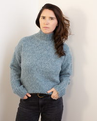 A woman with long brown hair wears the Hartford Myac Turtleneck Sweater in Jade and dark jeans, standing against a light wall with her hands in her pockets, looking seriously at the camera.