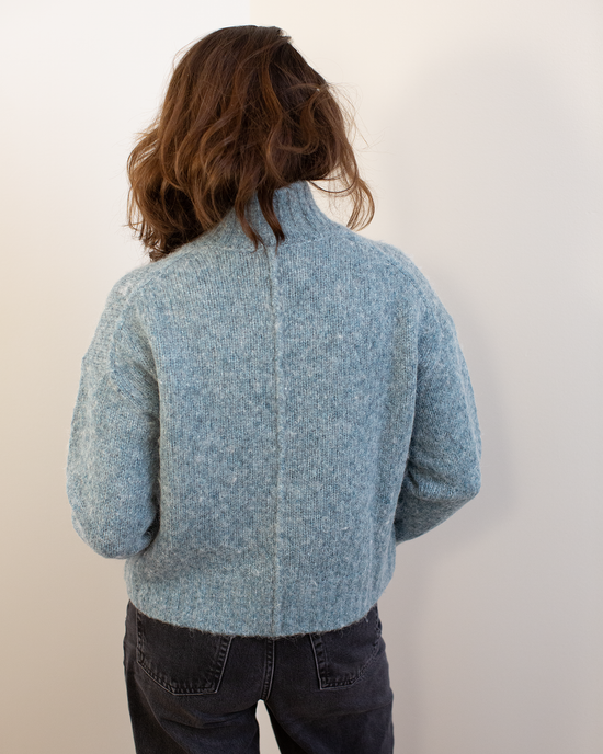 A person with wavy brown hair, wearing the Hartford Myac Turtleneck Sweater in Jade and dark jeans, stands facing away from the camera against a plain white wall.