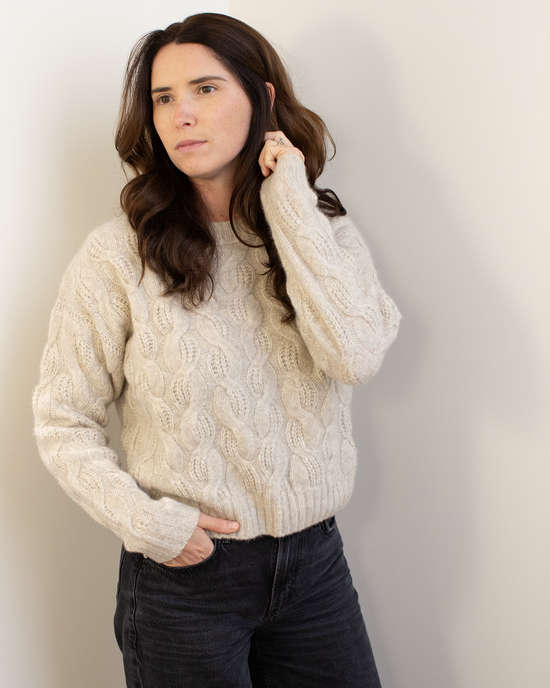 A woman with long brown hair wears the Autumn Cashmere Cable Crew in Sugarcane, styled in a relaxed fit with dark jeans. She stands against a light wall, one hand in her pocket and the other touching her hair, looking thoughtfully to the side.