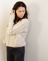 A woman with long brown hair wears the Autumn Cashmere Cable Crew in Sugarcane and black jeans, standing against a light wall and looking down with one hand in her hair.