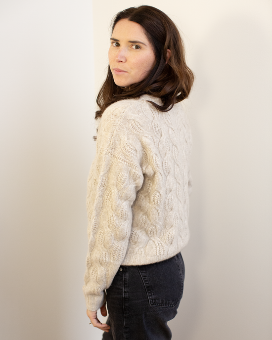 A woman with long brown hair models the Autumn Cashmere Cable Crew in Sugarcane, a relaxed fit cream-colored sweater, paired with dark jeans as she stands sideways and looks over her shoulder at the camera against a light background.