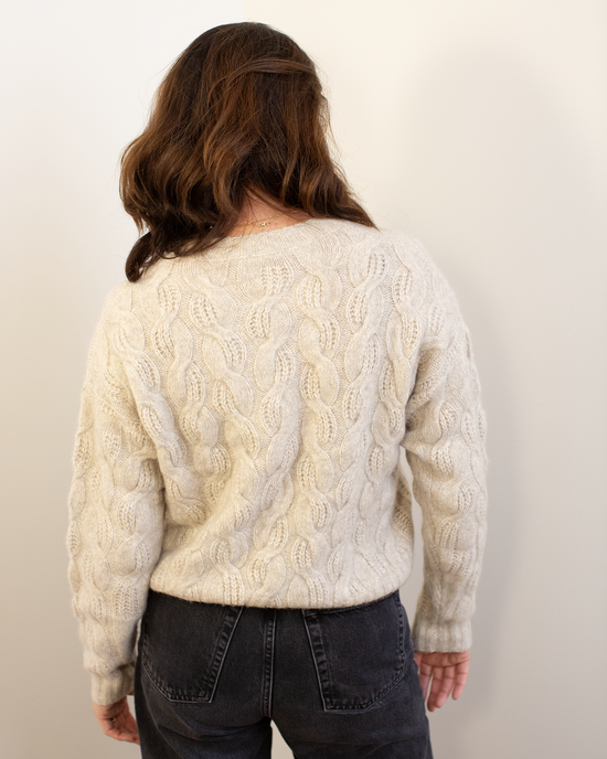 A person with wavy brown hair faces away, wearing the Autumn Cashmere Cable Crew in Sugarcane and high-waisted dark jeans against a plain light background.
