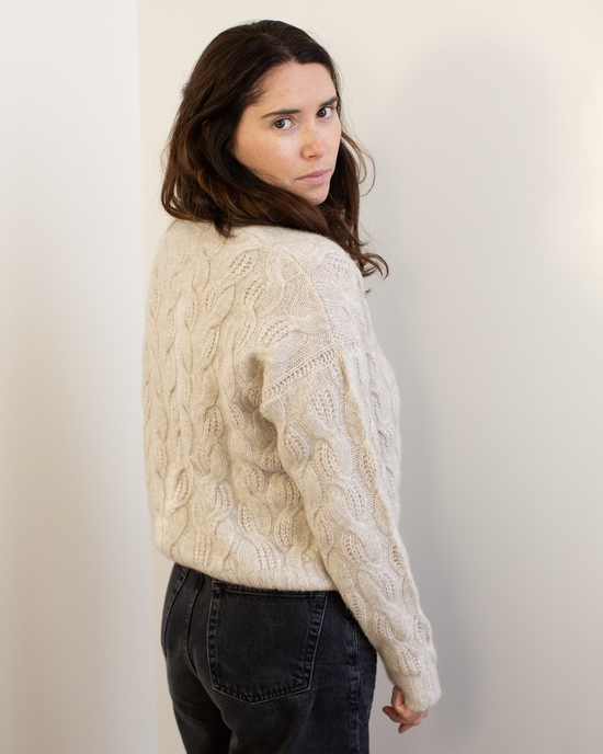 A woman with long brown hair stands sideways against a light wall, looking over her shoulder while wearing the Autumn Cashmere Cable Crew in Sugarcane and black jeans.