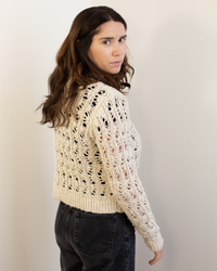 A woman with long brown hair wears the Autumn Cashmere Boxy Stitchery Cardigan in Oatmeal and dark jeans, standing before a plain light background. She looks over her shoulder at the camera with a neutral expression.