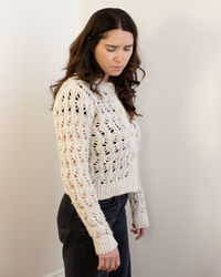 A woman with long brown hair wears the Autumn Cashmere Boxy Stitchery Cardigan in Oatmeal and dark pants, standing indoors against a plain light wall and looking down with a neutral expression.