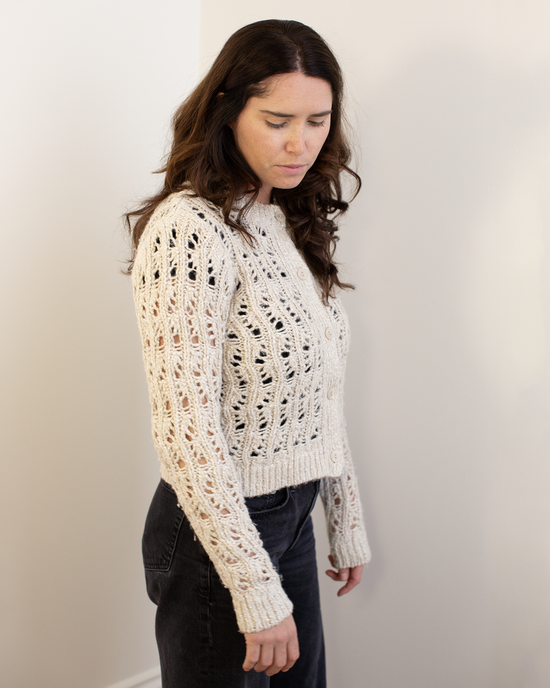 A woman with long brown hair wears the Autumn Cashmere Boxy Stitchery Cardigan in Oatmeal and dark pants, standing indoors against a plain light wall and looking down with a neutral expression.