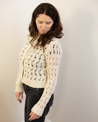 A woman with long brown hair wears the Autumn Cashmere Boxy Stitchery Cardigan in Oatmeal and black pants, standing sideways against a plain light background, looking down with a slight smile.