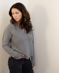 A woman with long brown hair wears the Autumn Cashmere Cable Sleeve Henley in Cement, paired with black pants, as she stands against a light wall looking down thoughtfully.