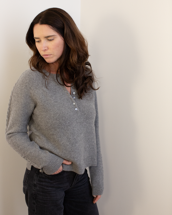A woman with long brown hair wears the Autumn Cashmere Cable Sleeve Henley in Cement, paired with black pants, as she stands against a light wall looking down thoughtfully.