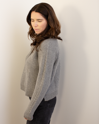 A woman with long brown hair stands in profile against a light wall, wearing the Autumn Cashmere Cable Sleeve Henley in Cement and dark jeans. She looks down with a neutral expression.