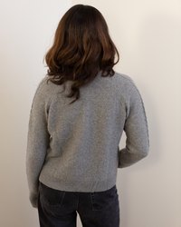 A person with wavy brown hair stands with their back to the camera, wearing the Autumn Cashmere Cable Sleeve Henley in Cement and dark gray jeans against a plain light-colored wall.