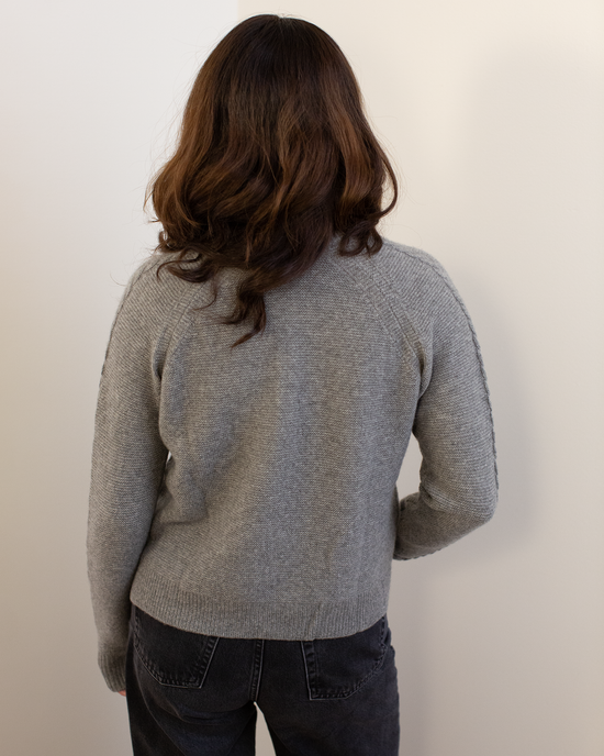 A person with wavy brown hair stands with their back to the camera, wearing the Autumn Cashmere Cable Sleeve Henley in Cement and dark gray jeans against a plain light-colored wall.