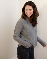 A woman with long brown hair, wearing the Autumn Cashmere Cable Sleeve Henley in Cement and dark jeans, stands against a light wall, smiling with her eyes closed and one hand in her pocket.
