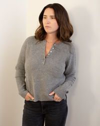 A woman with long brown hair models the Autumn Cashmere Cable Sleeve Henley in Cement, standing against a light wall with her hands in her pockets, wearing black pants and gazing slightly aside with a neutral expression.