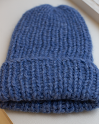A close-up of the Maiami Chunky Alpaca Hat in Blue Melange, handknit from soft alpaca with a folded brim, displayed on a light surface near a window.