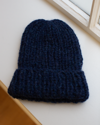 The Maiami Chunky Alpaca Hat in Denim, handknit and thick with a folded brim, rests on a white windowsill near a window.