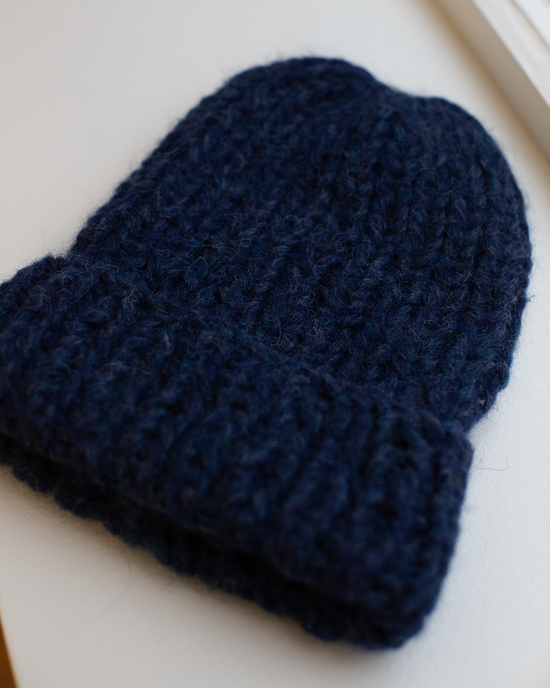The Maiami Chunky Alpaca Hat in Denim, a dark blue handknit beanie with a thick alpaca texture and folded brim, is displayed flat on a light surface by the window.