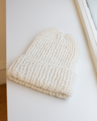 The Chunky Alpaca Hat in Creme by Maiami, handknit from a soft alpaca blend with a folded brim, rests on a white surface beside a window.