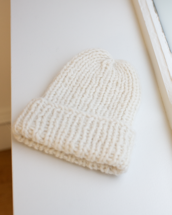 The Chunky Alpaca Hat in Creme by Maiami, handknit from a soft alpaca blend with a folded brim, rests on a white surface beside a window.
