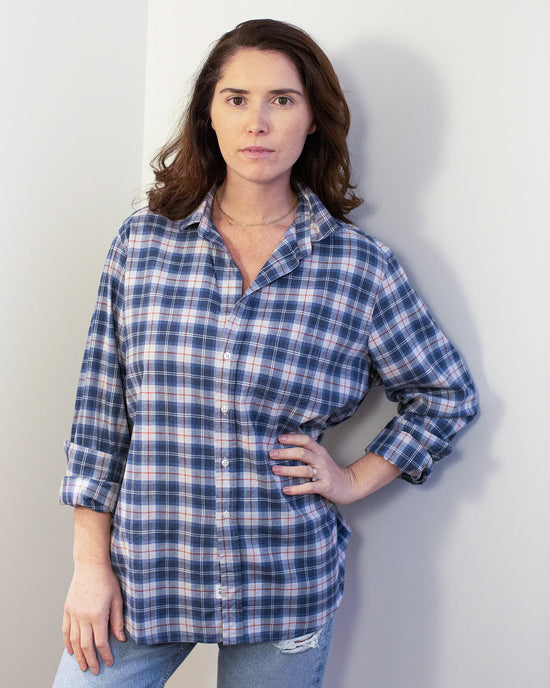 A woman with medium-length brown hair stands against a light wall, wearing the Frank & Eileen Eileen Relaxed Button Up Shirt in Grey & Red Plaid and light blue ripped jeans, with one hand on her hip and a neutral expression.