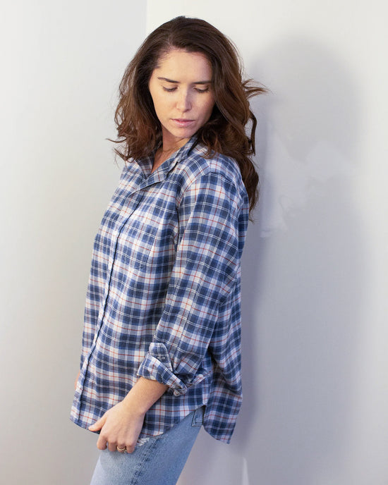A woman with wavy brown hair wears the Frank & Eileen Eileen Relaxed Button Up Shirt in Grey & Red Plaid and light blue jeans, standing against a white wall and looking down to her left with a calm expression.