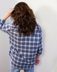 A person with long, wavy brown hair faces away from the camera, wearing the Frank & Eileen Eileen Relaxed Button Up Shirt in Grey & Red Plaid and light jeans, with one hand raised to their head.