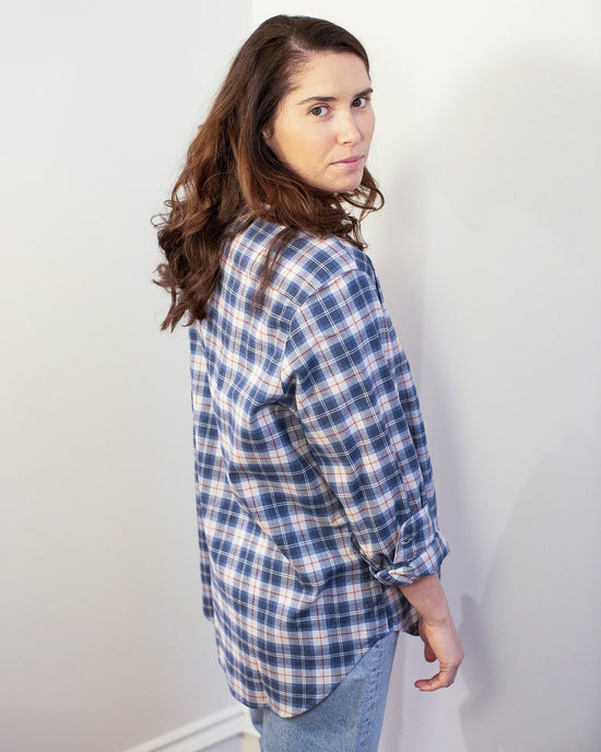 A woman with long brown hair stands sideways against a plain light wall, wearing the Frank & Eileen Eileen Relaxed Button Up Shirt in Grey & Red Plaid with jeans, looking back over her shoulder.