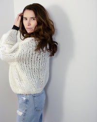A woman with long brown hair wears the Maiami Mohair Bomber Cardigan in Creme, standing sideways near a white wall in light blue ripped jeans and looking at the camera with a neutral expression.