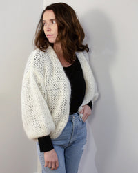 A woman with wavy brown hair wears the Maiami Mohair Bomber Cardigan in Creme over a black top and light blue jeans, standing against a plain light-colored wall and looking to her left.