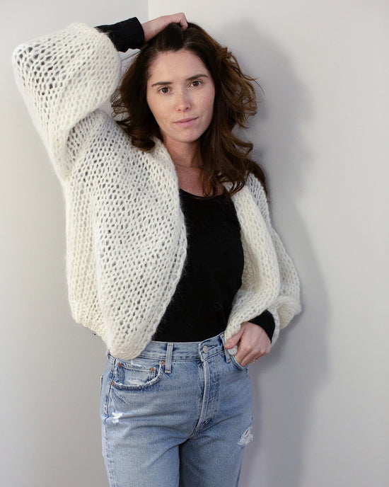 A woman with long brown hair wears the Maiami Mohair Bomber Cardigan in Creme over a black top and light blue ripped jeans. She stands against a plain light wall, one hand in her pocket, the other touching her hair.