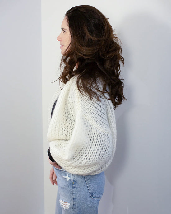 A woman with long, wavy brown hair stands in profile against a white wall, wearing the Maiami Mohair Bomber Cardigan in Creme over a black top and light blue ripped jeans.