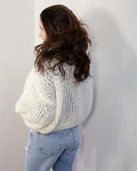 A person with long, wavy brown hair stands sideways facing a white wall, wearing the Maiami Mohair Bomber Cardigan in Creme and light blue jeans.