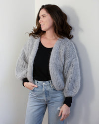 A woman with long brown hair smiles with one hand in her pocket, wearing Maiami’s oversized Mohair Bomber Cardigan in Concrete over a black shirt and light blue jeans, standing against a white wall.