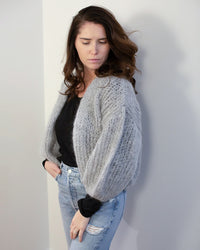 A woman with long brown hair wears the Maiami Mohair Bomber Cardigan in Concrete over a black top and light blue ripped jeans, standing against a white wall and looking down with one hand in her pocket.