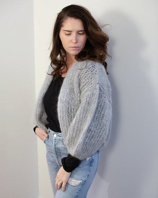 A woman with long brown hair wears the Maiami Mohair Bomber Cardigan in Concrete over a black top and light blue ripped jeans, standing against a white wall and looking down with one hand in her pocket.