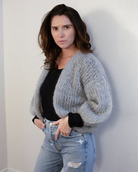A woman with wavy brown hair wears the Maiami Mohair Bomber Cardigan in Concrete over a black top and light blue ripped jeans, standing confidently against a plain, light-colored wall and looking at the camera.