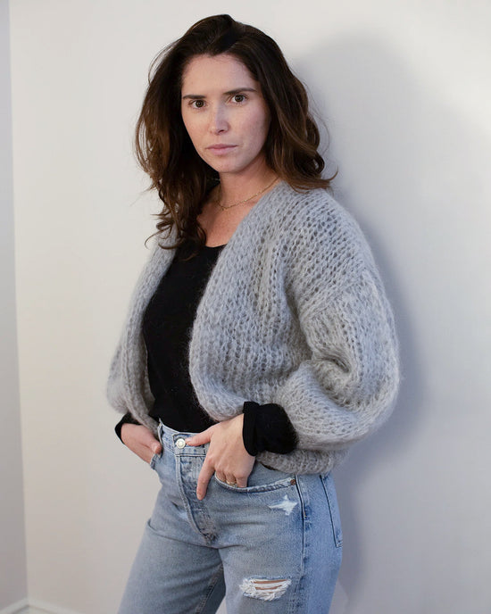 A woman with wavy brown hair wears the Maiami Mohair Bomber Cardigan in Concrete over a black top and light blue ripped jeans, standing confidently against a plain, light-colored wall and looking at the camera.