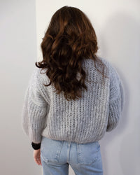 A person with long, wavy brown hair stands with their back to the camera, wearing the Maiami Mohair Bomber Cardigan in Concrete and light blue jeans against a plain, light-colored wall.