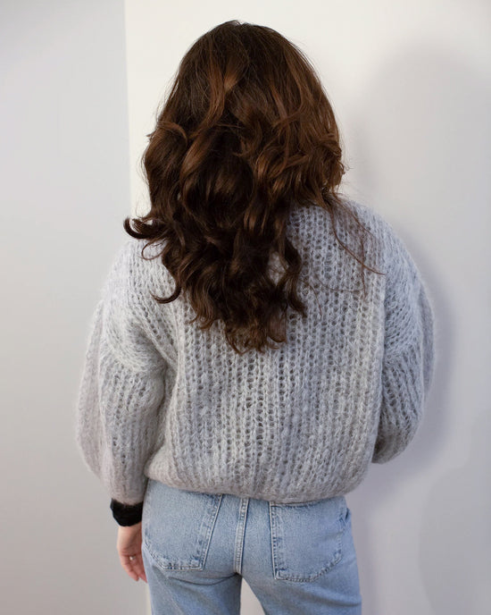 A person with long, wavy brown hair stands with their back to the camera, wearing the Maiami Mohair Bomber Cardigan in Concrete and light blue jeans against a plain, light-colored wall.