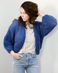 A woman with long brown hair wears the Maiami Mohair Bomber Cardigan in Dark Blue over a light beige t-shirt and light blue ripped jeans, looking down and touching her hair against a plain background.