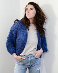 A woman with long brown hair wears the Maiami Mohair Bomber Cardigan in Dark Blue over a light t-shirt and ripped jeans, standing against a white wall with her hands in her pockets and gazing thoughtfully to the side.
