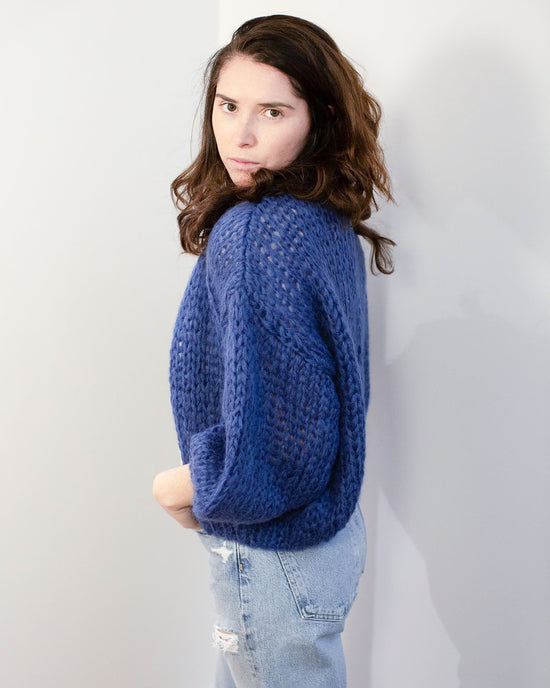 A person with long brown hair wears the Maiami Mohair Bomber Cardigan in Dark Blue and light-wash ripped jeans, standing sideways against a plain white wall and looking directly at the camera with a neutral expression.