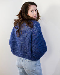 A woman with long brown hair wears the Maiami Mohair Bomber Cardigan in Dark Blue and light blue jeans, standing against a plain white background and looking over her shoulder at the camera.