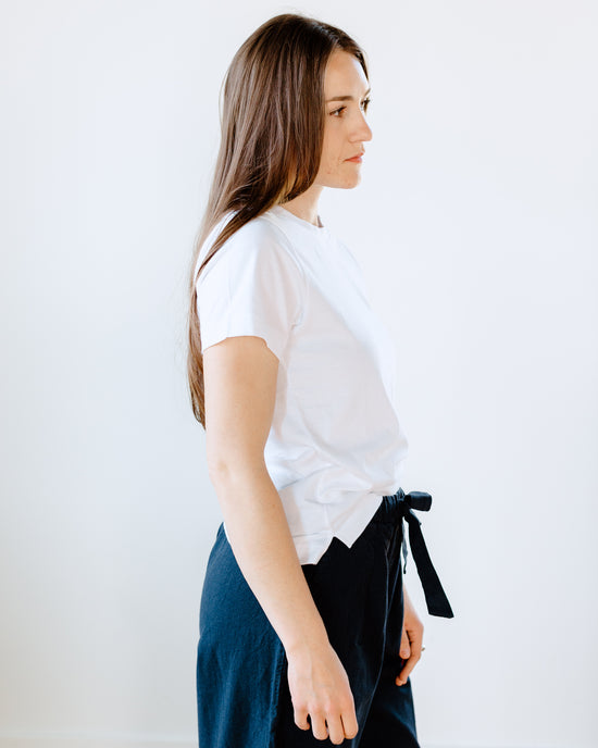 Woman standing in profile wearing a classic fit crew neck Maliah Organic Cotton T-Shirt in White by Beaumont Organic and dark trousers with a tied waistband.