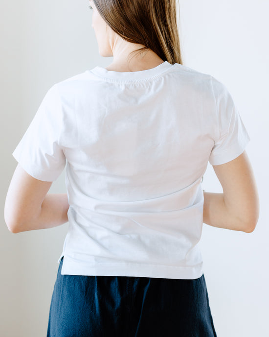 A person from behind wearing a Maliah Organic Cotton T-Shirt in White by Beaumont Organic and dark trousers against a light background.