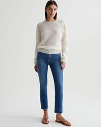 A woman standing in a white studio, wearing a cream sweater and medium indigo blue straight leg jeans, paired with brown sandals from AG Jeans' Mari Crop in Alibi Destructed.