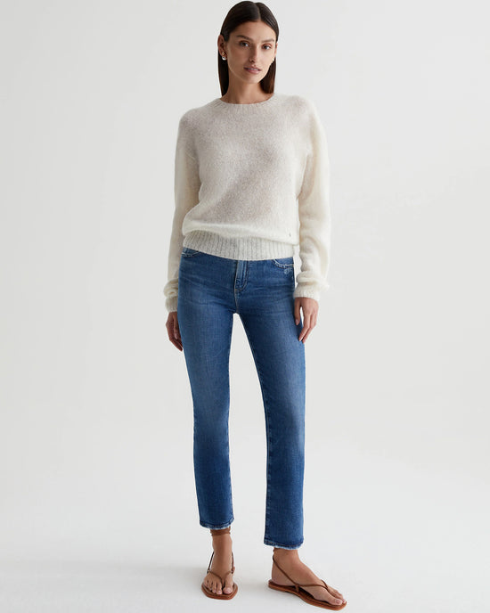 A woman standing in a white studio, wearing a cream sweater and medium indigo blue straight leg jeans, paired with brown sandals from AG Jeans' Mari Crop in Alibi Destructed.