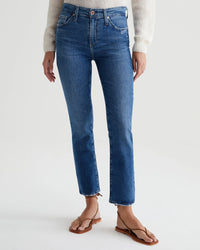 Woman standing in AG Jeans Mari Crop in Alibi Destructed fitted medium indigo blue straight leg jeans and brown sandals, with a white sweater partially visible at the top. Jeans have a frayed hem.